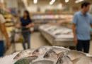 Shoppers walking past a supermarket seafood section with whole milkfish, tilapia, and parrotfish on ice, viewed at an angle with the display slightly blurred.