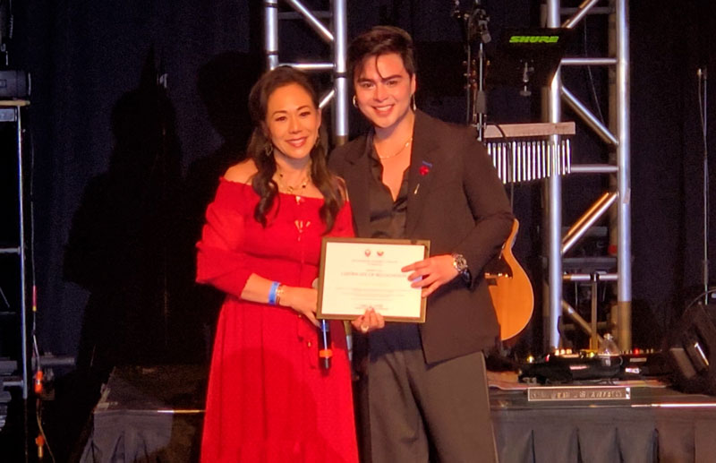 Honorary Consul Jenny Ho-Vijungco in a red dress stands alongside Mark Mabasa on stage at Casino Arizona, both holding a framed certificate of recognition from the Philippine Honorary Consulate of Arizona.